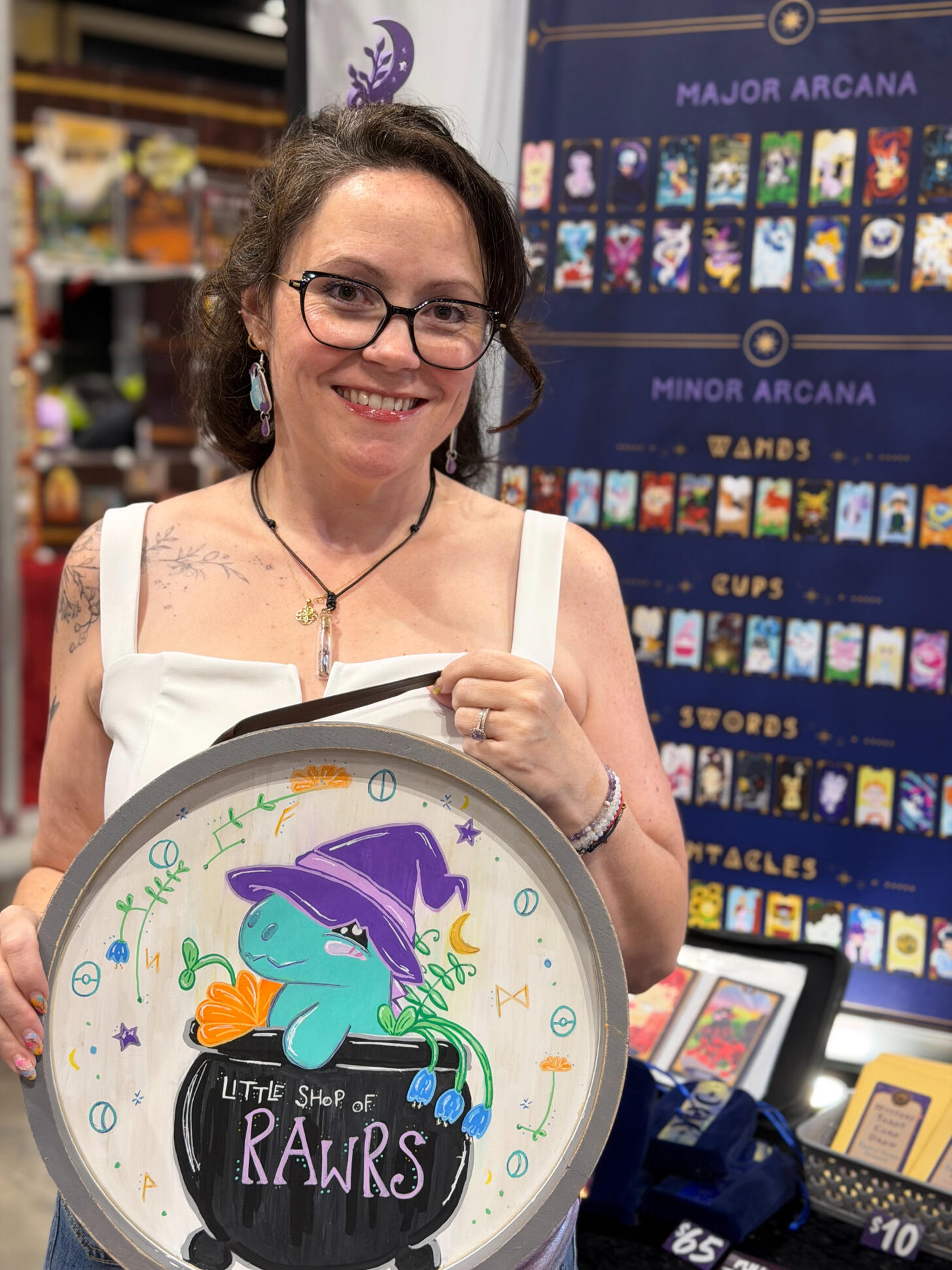 Heather smiling at her Little Shop of RAWRS convention booth, surrounded by Pokemon art prints and stickers